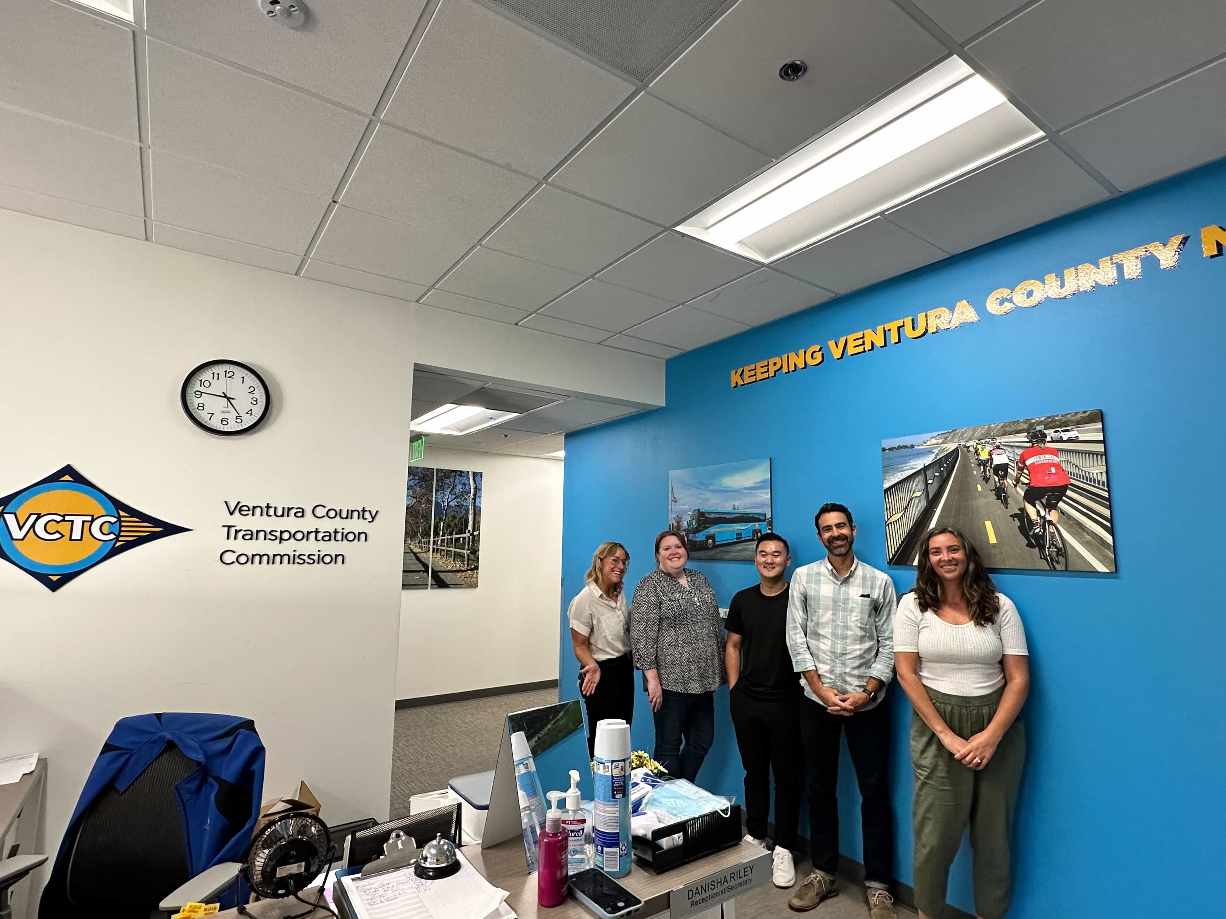 Five members of the VCTC team pose in their office in front of a blue wall with the text "Keeping Ventura County Moving."